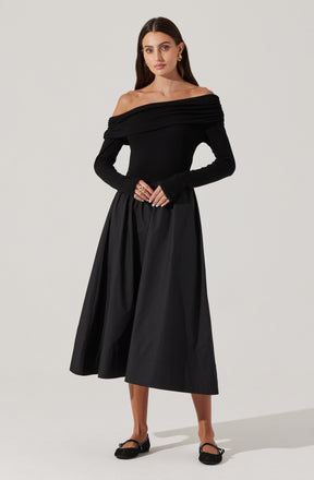 A woman is wearing an off-the-shoulder black top with a wide, ruched neckline that drapes across her shoulders. She pairs it with a high-waisted, full-length black skirt that has a slightly shiny, textured fabric. The outfit is accessorized with earrings and rings, and she is wearing black flats.