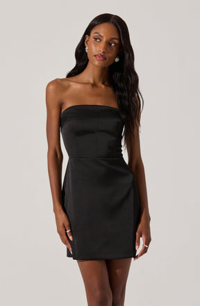 A woman is wearing a strapless black dress that is fitted at the bodice and falls above the knees. The dress has a smooth, shiny fabric appearance. She has long, wavy black hair and is accessorized with large, circular earrings and rings on her fingers.