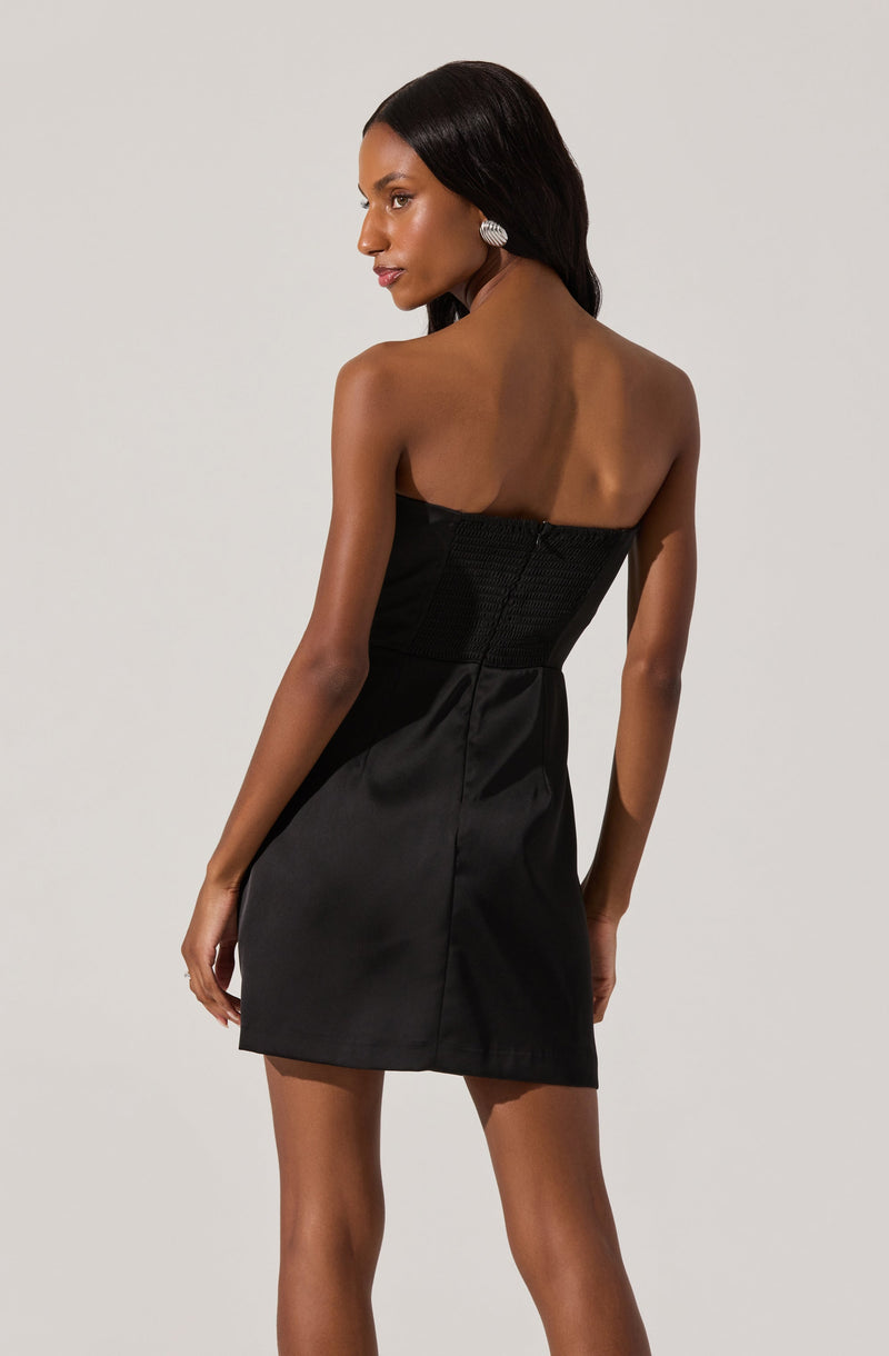 A woman is wearing a strapless black dress that features a fitted, textured bodice and a smooth, slightly shiny skirt. The dress is knee-length, and she accessorizes with large, round, silver earrings.