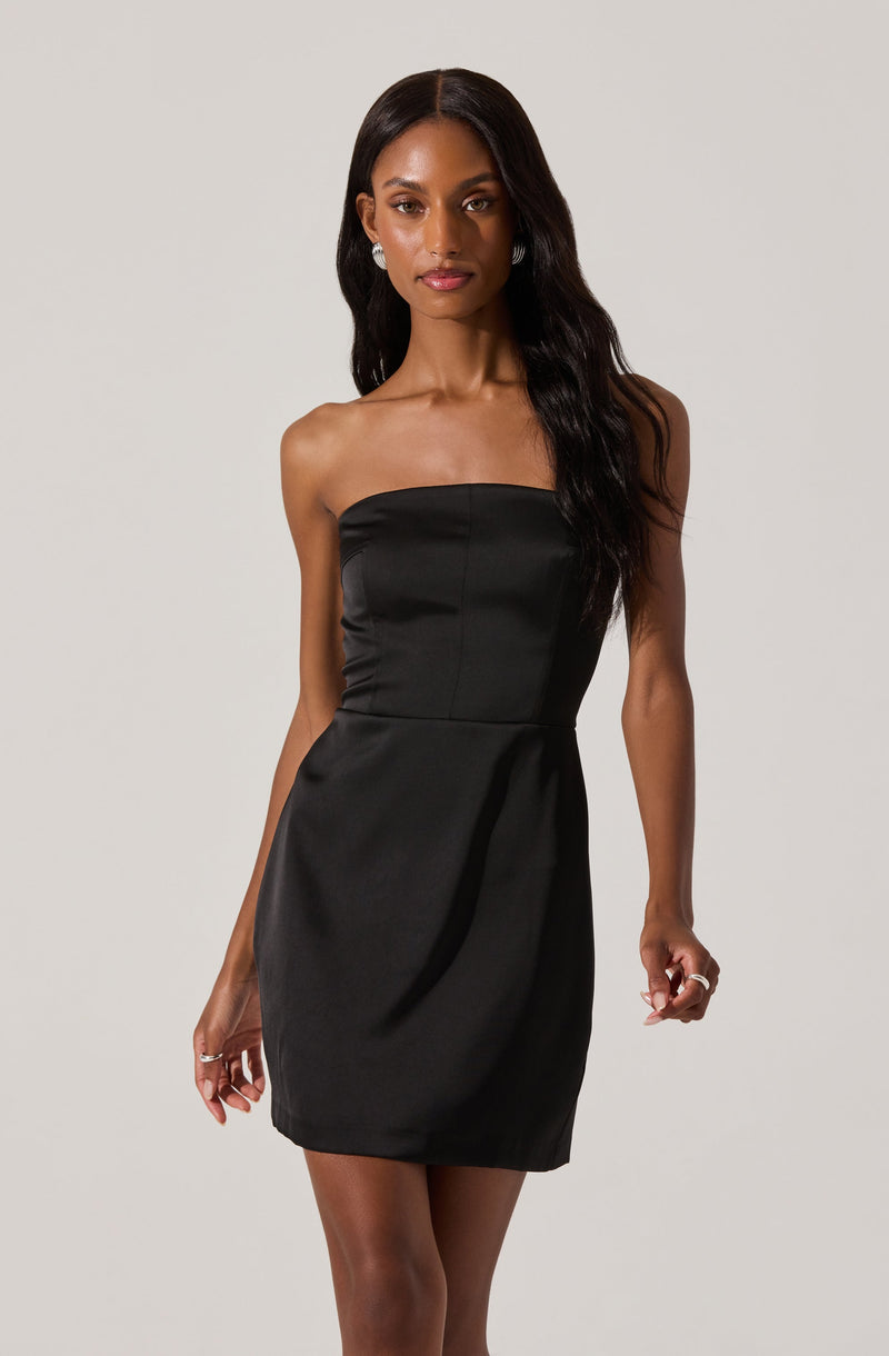 A woman is wearing a strapless black dress that is fitted at the bodice and has a slightly gathered skirt. The dress is smooth and appears to be made of a shiny fabric. She is accessorized with small earrings and rings.