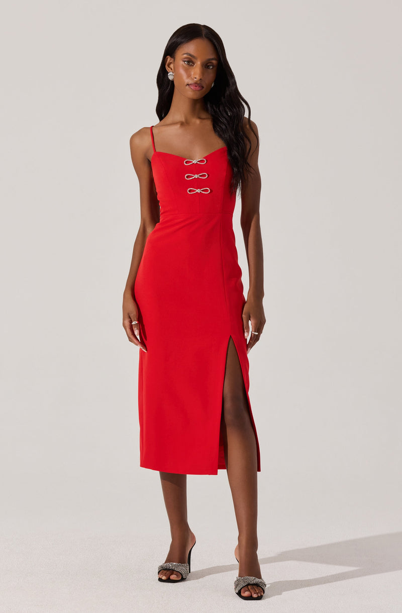 A woman is wearing a fitted red midi dress with thin spaghetti straps. The dress features three decorative bow-shaped buttons on the front bodice and a high slit on the left side. She is also wearing open-toe high-heeled shoes with embellishments and earrings.