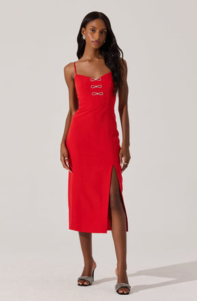 A woman is wearing a fitted red midi dress with thin spaghetti straps. The dress features three decorative bow-shaped buttons on the front bodice and a high slit on the left side. She is also wearing open-toe high-heeled shoes with embellishments and earrings.