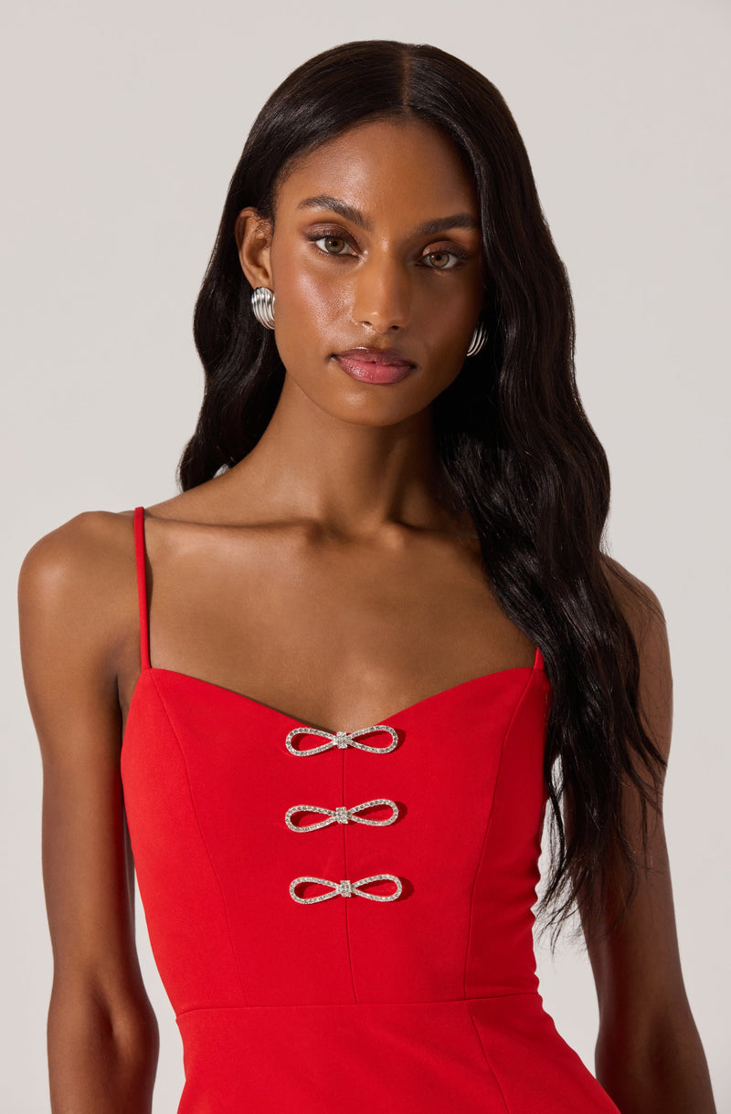 A woman is wearing a sleeveless, fitted red dress with thin spaghetti straps. The dress features three decorative bows in a silver, rhinestone-encrusted design down the center front.