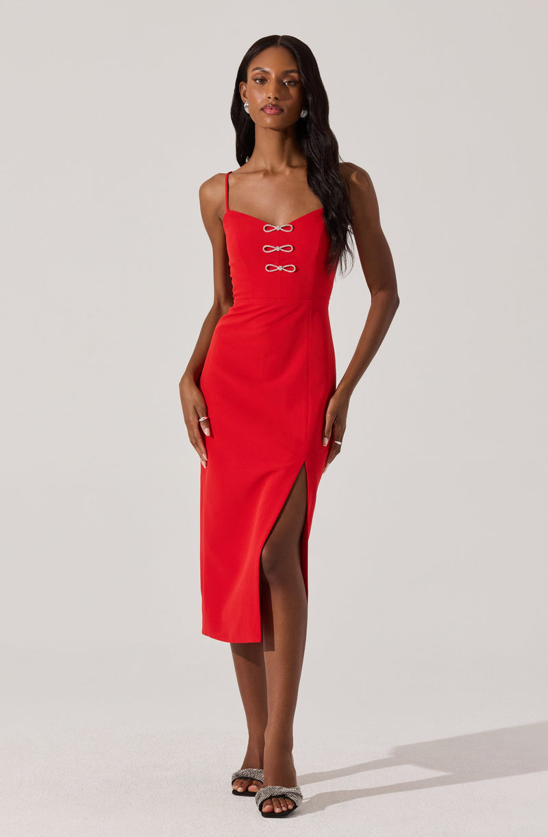 A woman is wearing a fitted, sleeveless red dress with spaghetti straps. The dress features a decorative detail with three small bows on the front and has a high slit on one side, revealing part of her leg. She is also wearing open-toe high-heeled shoes with embellishments.