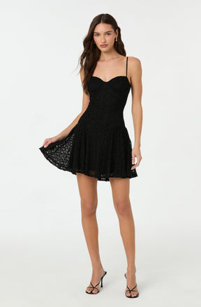 A young woman is wearing a black lace dress with a fitted bodice featuring a sweetheart neckline and thin spaghetti straps. The dress has a flared, above-the-knee skirt with a scalloped lace hem. She is also wearing black strappy high-heeled sandals.