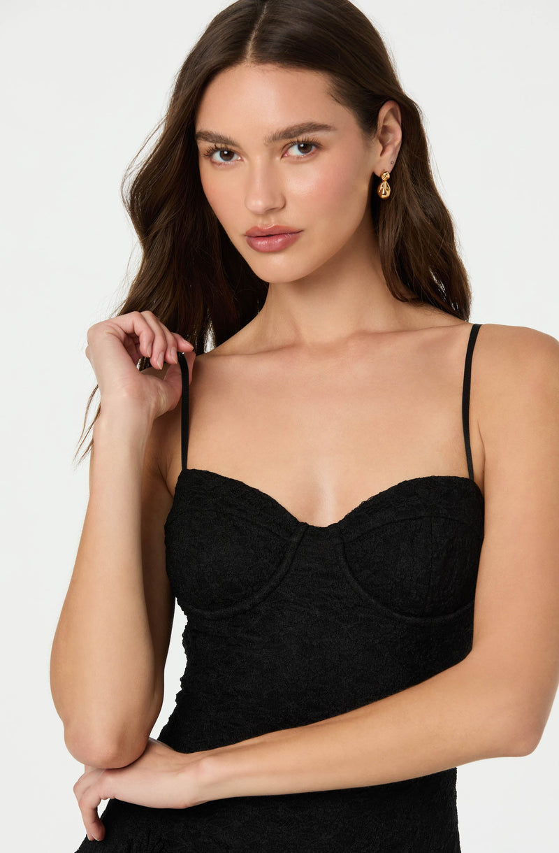 A woman is wearing a black, textured dress with thin spaghetti straps and a fitted bodice. She has gold earrings and her long brown hair is styled in loose waves.