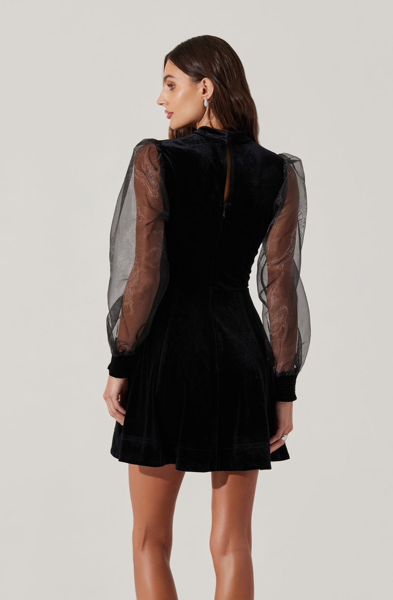 A woman is wearing a black dress with a velvet bodice and sheer, puffed long sleeves made of transparent fabric. The dress has a high neckline and a keyhole opening at the back with a button closure. The skirt of the dress is short and slightly flared.