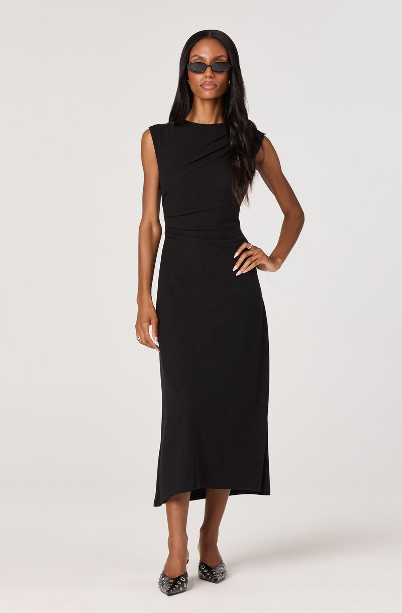 A woman is wearing a sleeveless, black, ankle-length dress with a high neckline and subtle draping details on the bodice. She pairs the dress with black sunglasses and pointed-toe heels with a black and white snakeskin pattern.
