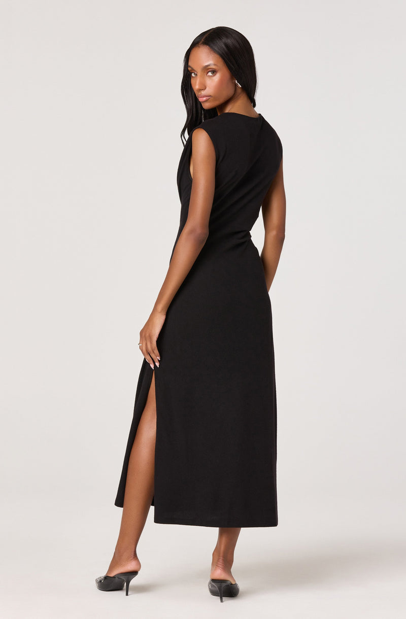 A woman is wearing a sleeveless, long black dress with a slit on the side, revealing her leg. The dress is fitted and ankle-length, paired with black high-heeled shoes.
