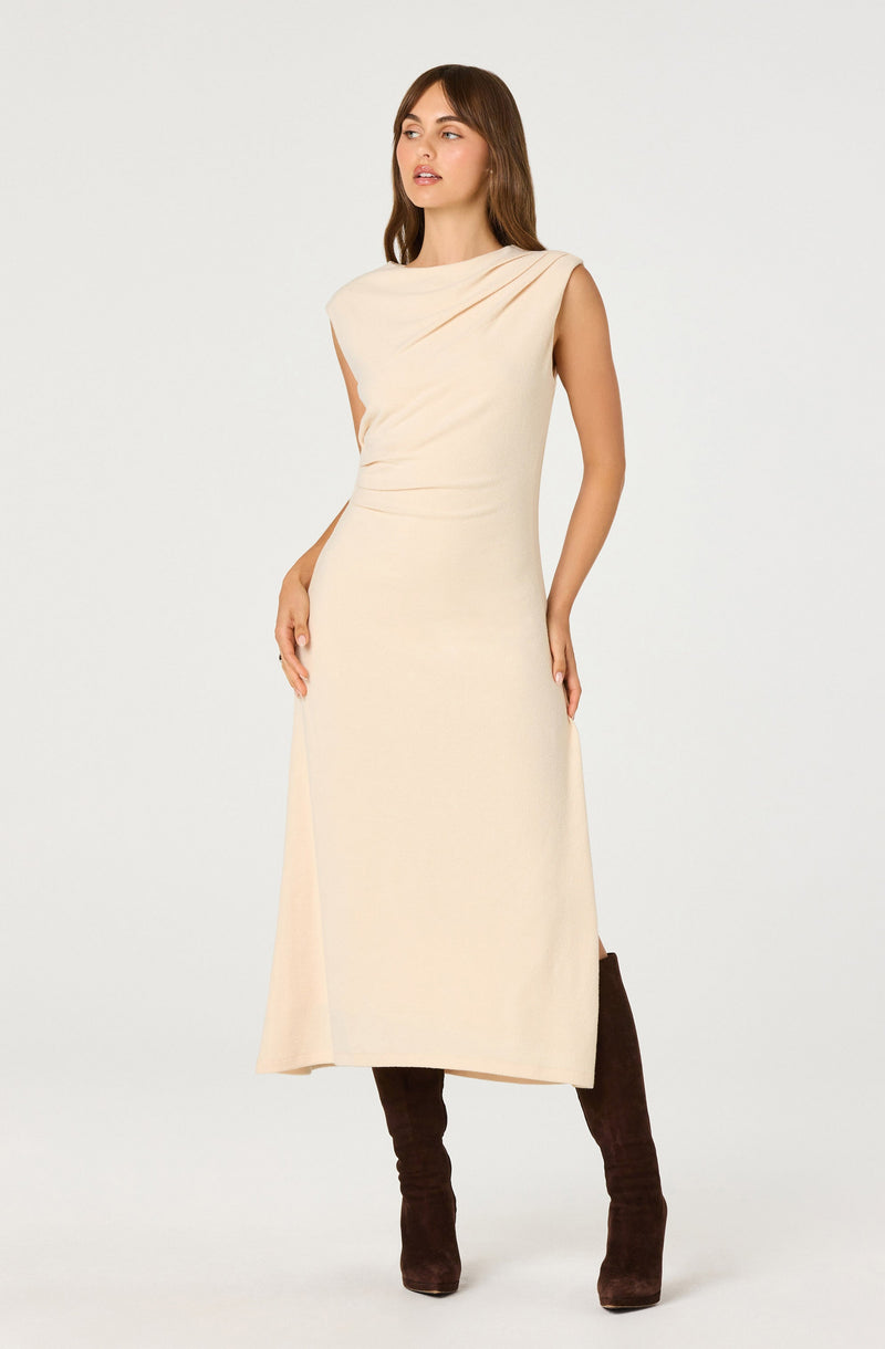 A woman is wearing a sleeveless, light beige, ankle-length dress with a high neckline and slight draping details on the shoulder and chest area. She has paired the dress with dark brown, knee-high boots.