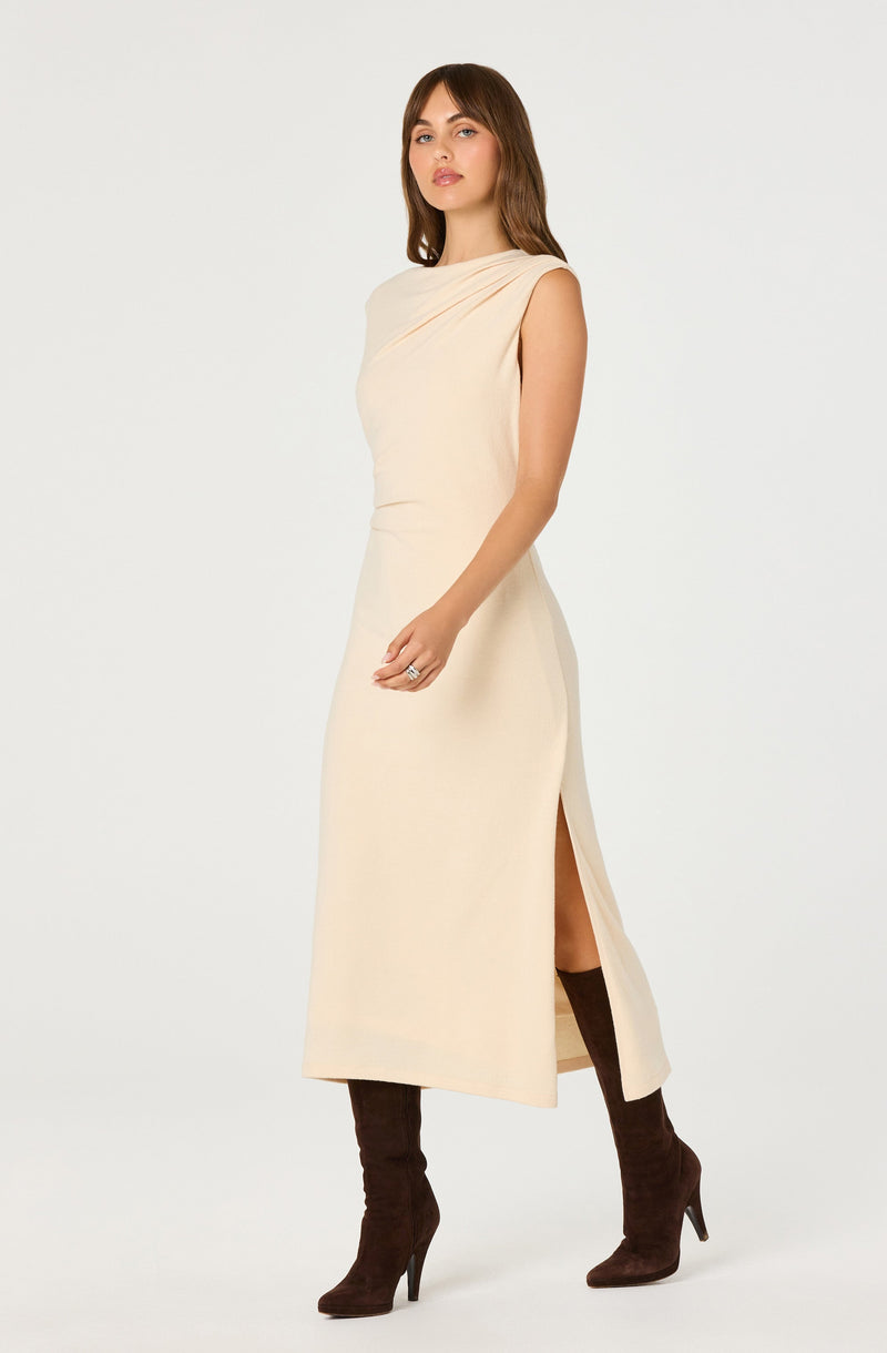 A woman is wearing a sleeveless, knee-length, creamy beige dress with a draped neckline and a side slit. She has on dark brown high-heeled boots.