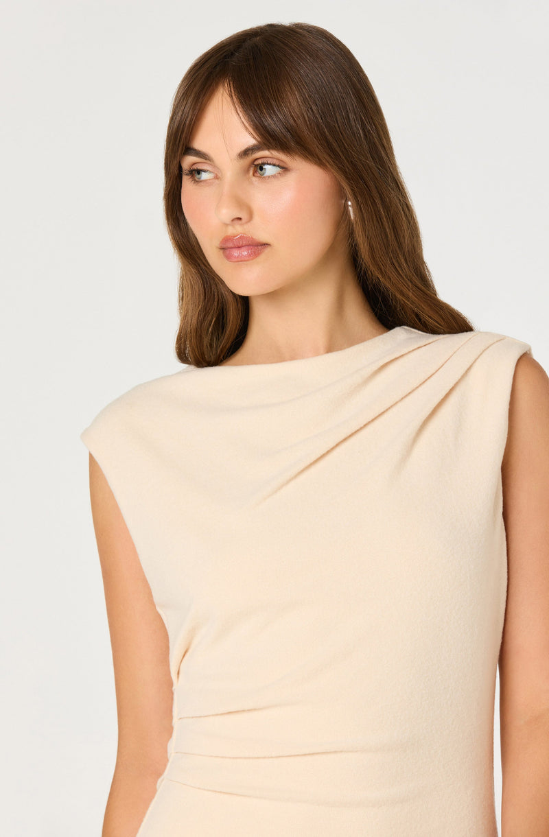 A woman with shoulder-length brown hair is wearing a sleeveless, cream-colored dress with a draped, cowl neckline.