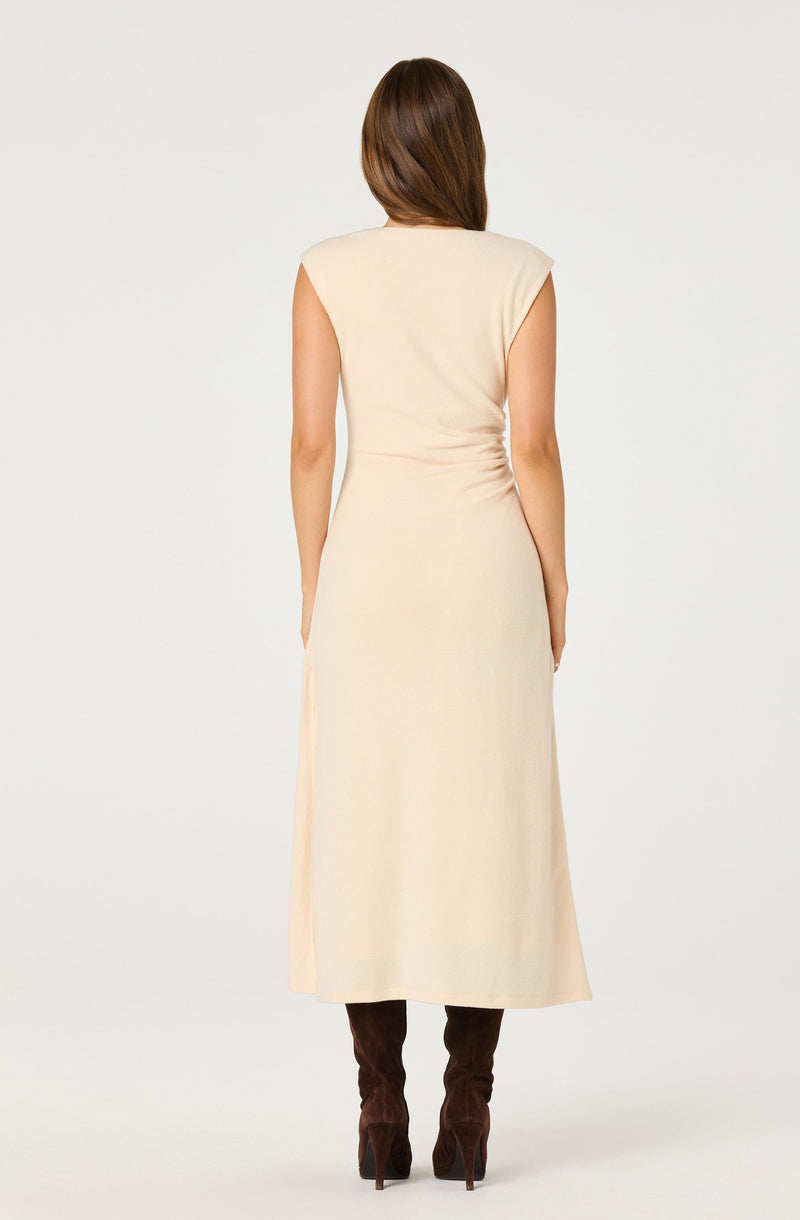 A woman is wearing a sleeveless, knee-length, cream-colored dress with a loose fit at the top and a slight flare at the bottom. She has on brown, high-heeled boots.