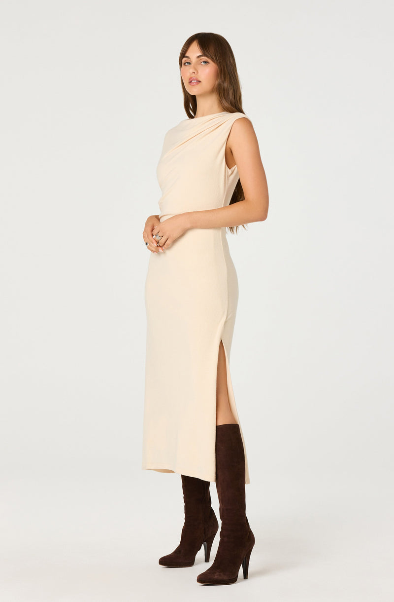 A woman is wearing a sleeveless, cream-colored, floor-length dress with a draped neckline and a side slit that extends to her knee. She has long, brown hair and is wearing dark brown high-heeled knee-high boots.