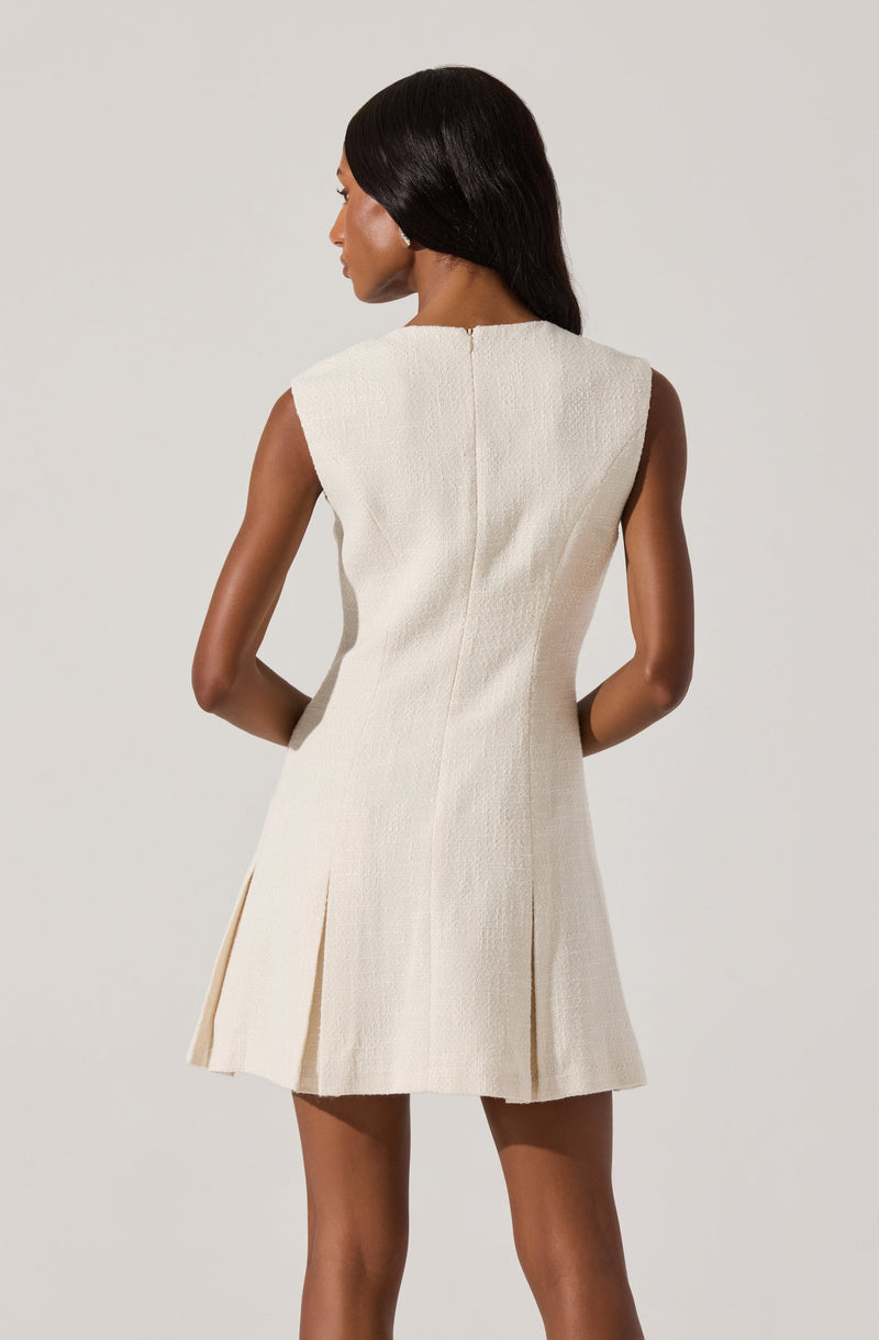 A woman is wearing a sleeveless, cream-colored textured dress with a straight neckline and a back zipper. The dress has a fitted waist and a pleated, slightly flared skirt that falls above the knees.