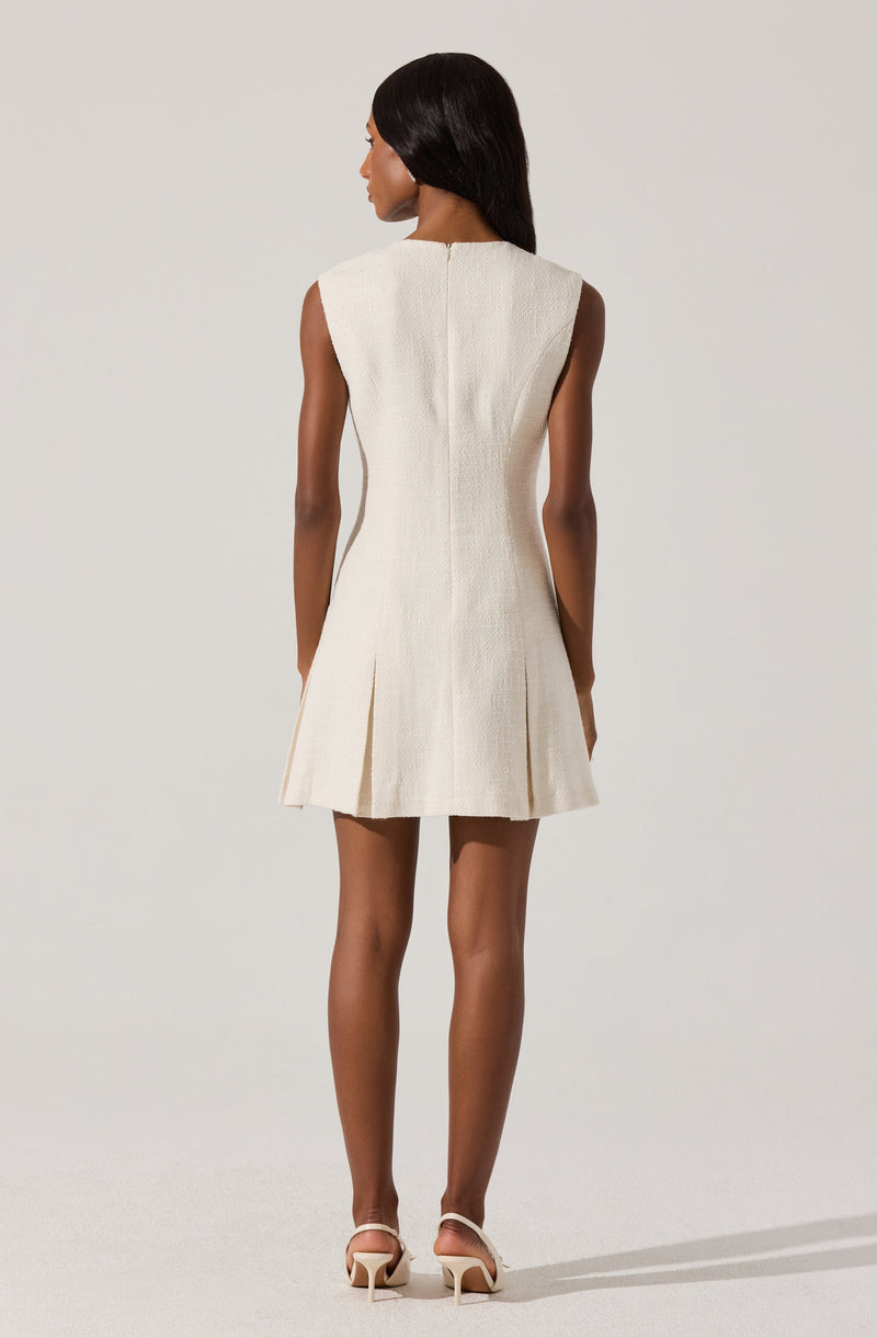 A woman is wearing a sleeveless, cream-colored, textured dress that is fitted through the waist and flares out into an A-line shape with subtle pleats at the hem. The dress has a clean, minimalist design with no visible embellishments, and she is paired with nude high-heeled shoes.