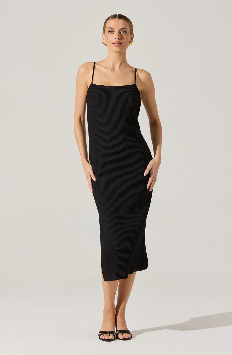 A woman is wearing a sleeveless black dress with thin spaghetti straps. The dress has a ribbed texture and falls to mid-calf length. She is wearing black open-toe sandals with thin straps.