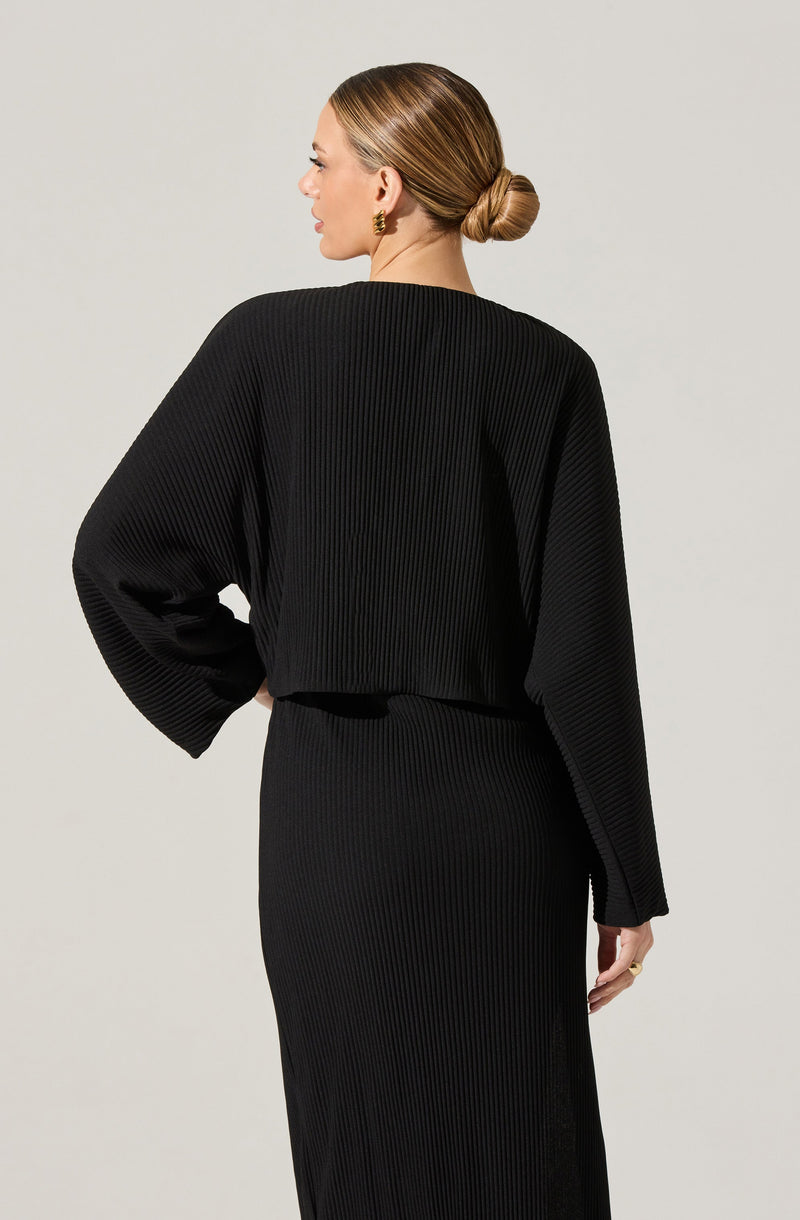 A woman is wearing a black, ribbed, long-sleeve top with a loose fit and wide sleeves. She pairs it with a matching black ribbed skirt that extends to the floor. Her hair is styled in a neat, low bun, and she is wearing gold earrings.