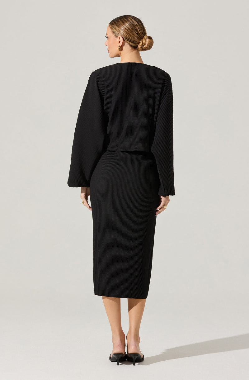 A woman is wearing a black, ribbed knit outfit consisting of a long-sleeved top with a round neckline and a matching high-waisted, knee-length pencil skirt. She is also wearing black high-heeled shoes.