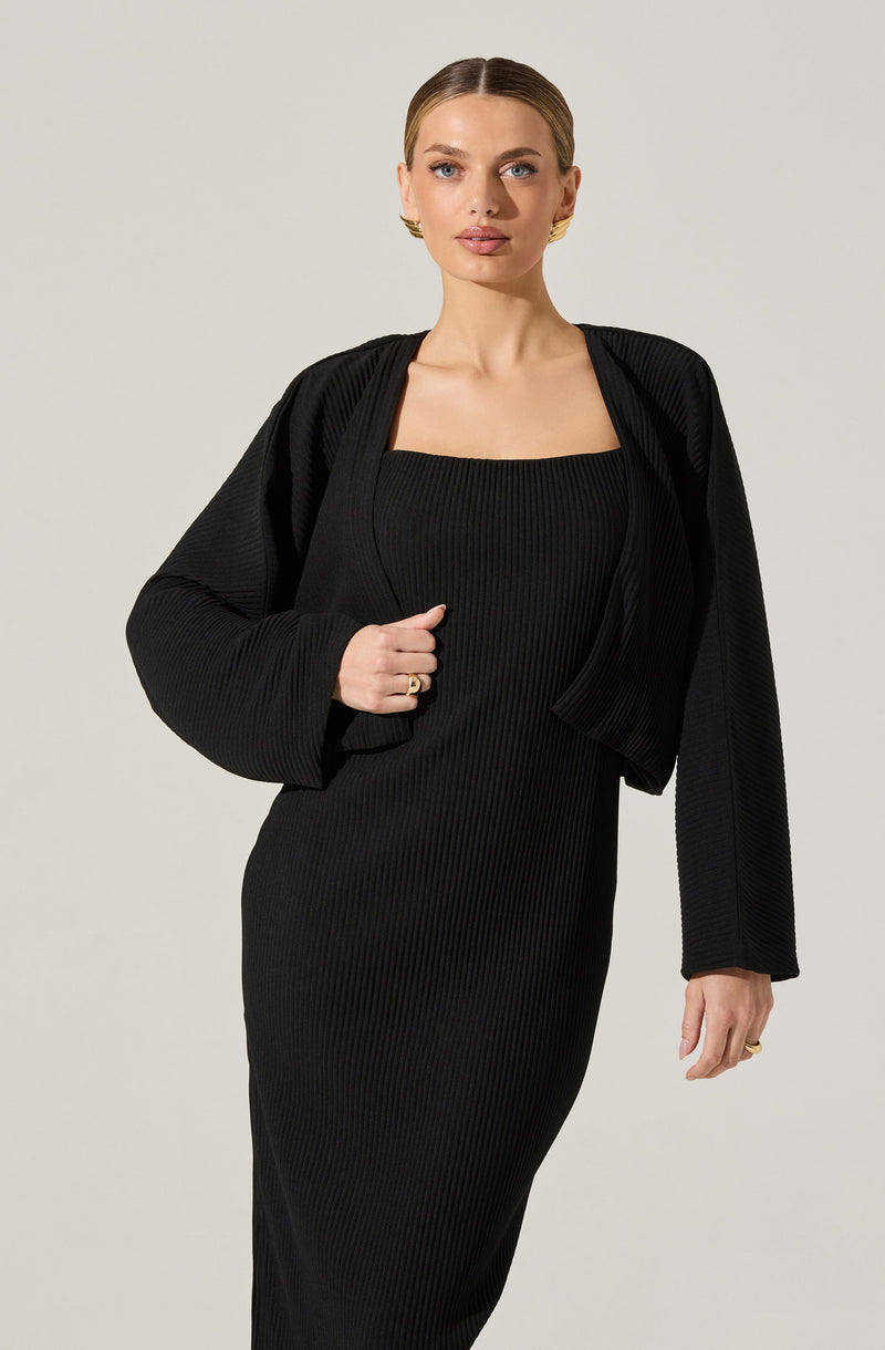 A woman is wearing a black, ribbed, fitted dress with a square neckline. She has a matching black, ribbed, cropped blazer draped over her shoulders.
