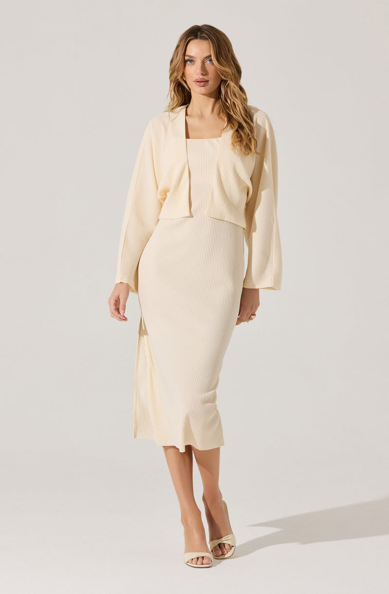 A woman is wearing a matching set in a light cream color. The outfit consists of a sleeveless, ribbed, square-neck top and a high-waisted, ribbed, midi-length skirt with a side slit. She has a loose, open-front cardigan draped over her shoulders with long sleeves. She is also wearing light-colored, open-toe high heels.