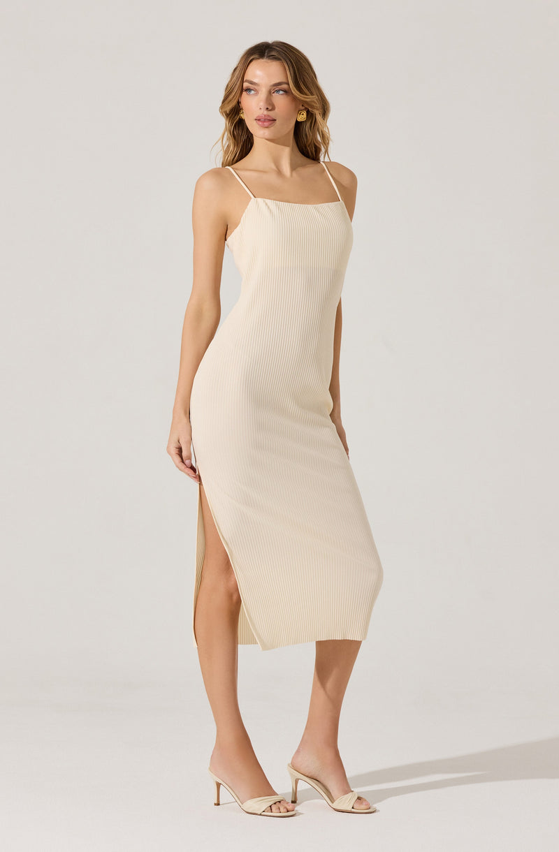 A woman is wearing a sleeveless, cream-colored midi dress with thin spaghetti straps. The dress features a ribbed texture and side slits up to the knee. She has paired the dress with open-toe heels that match the dress color and has gold earrings.