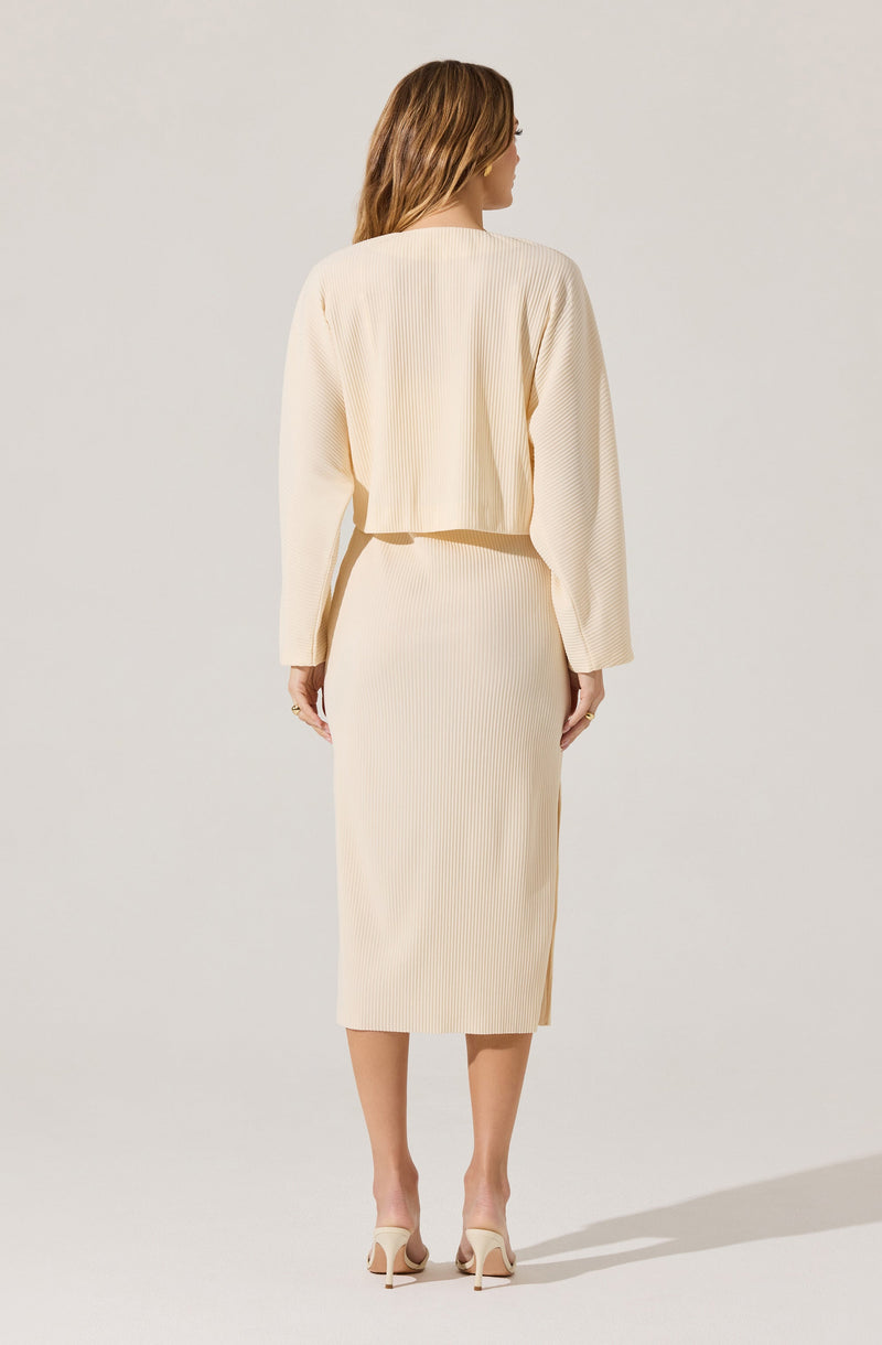 A woman is wearing a matching cream-colored outfit consisting of a cropped, long-sleeved, textured top and a high-waisted, ribbed midi skirt. She is also wearing light-colored, open-toe high heels. The overall style appears elegant and minimalistic.