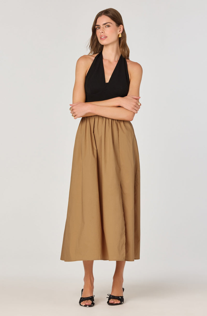 A woman is wearing a sleeveless black top with a V-neckline and wide shoulder straps. She pairs it with a high-waisted, full-length beige skirt that has an elastic waistband. She completes her outfit with black open-toed high-heeled sandals. She has long brown hair, gold earrings, and a relaxed pose with arms crossed.