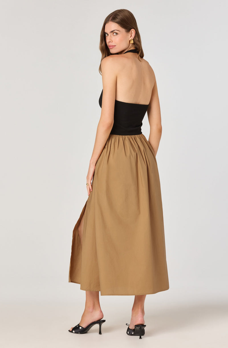 A woman is wearing a black strapless top that fits snugly around her torso and a tan, high-waisted, ankle-length skirt with a gathered waist and a slit on the side. She is also wearing black high-heeled mules with bow accents.