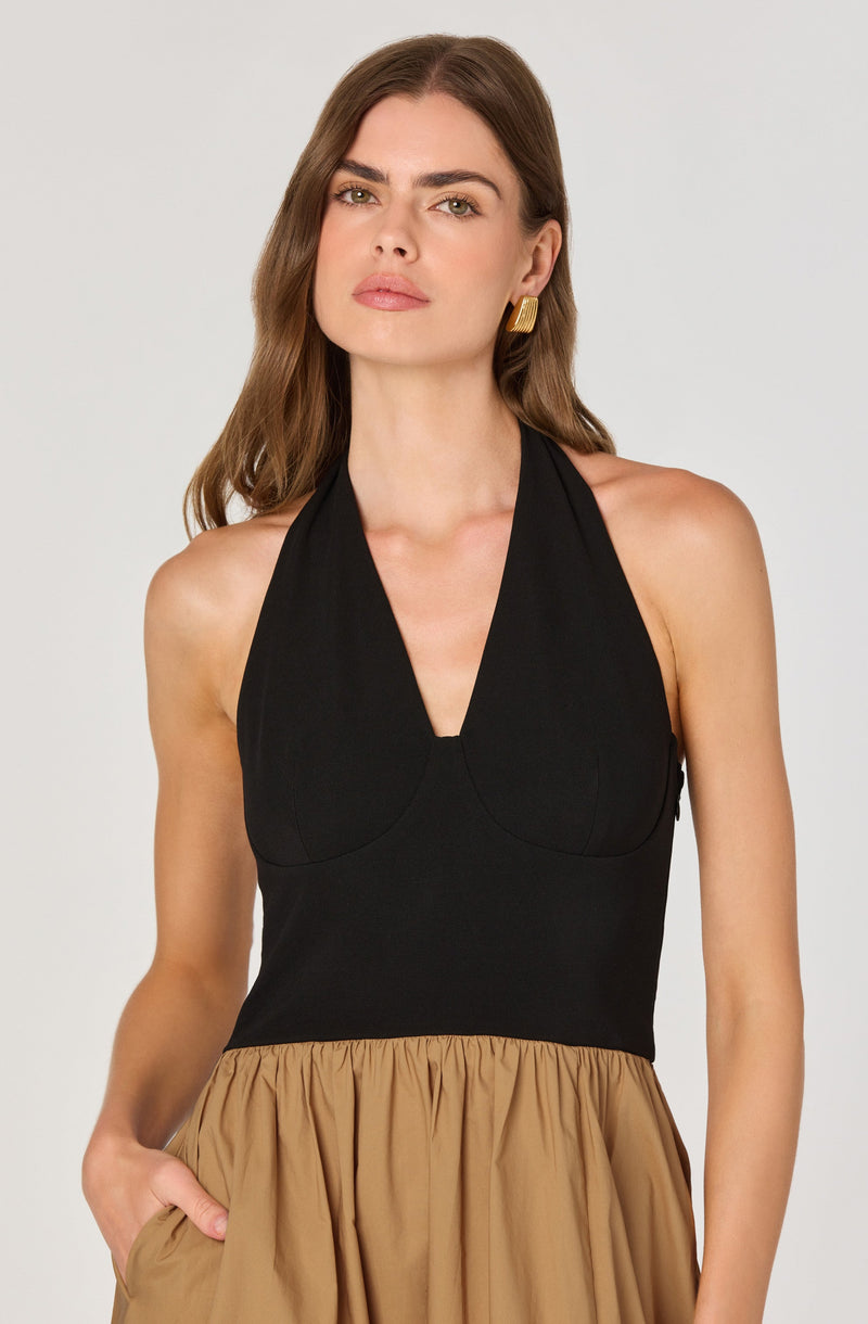 A woman is wearing a sleeveless, halter-neck black top with a fitted waist, paired with a high-waisted tan skirt that has a gathered, flowing design. She accessorizes with large, rectangular, gold earrings.