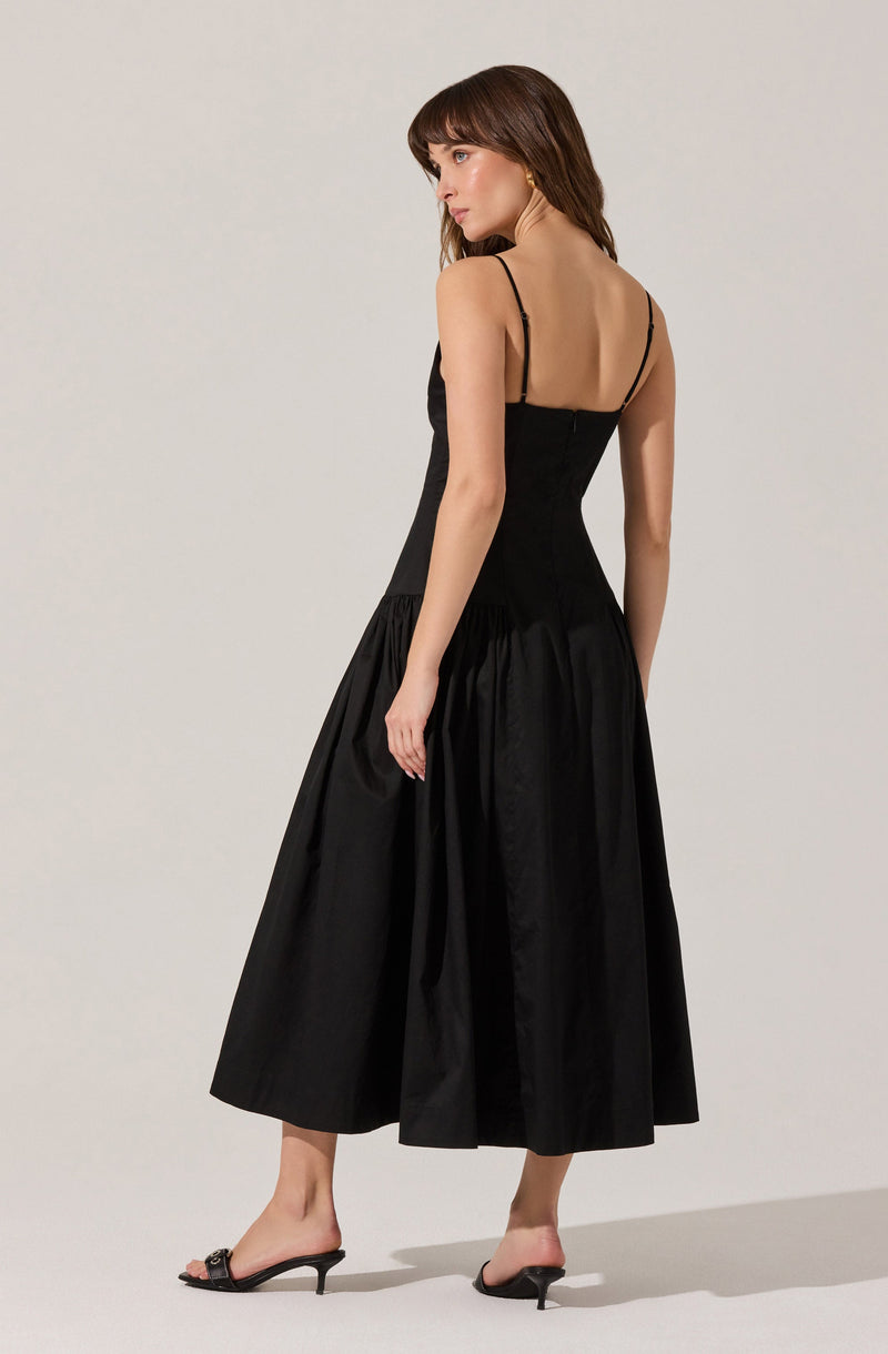 A woman is wearing a black dress with thin spaghetti straps, a fitted bodice, and a high waist. The skirt is long, full, and gathered, creating a voluminous and elegant appearance. She is also wearing black open-toe high-heeled sandals.
