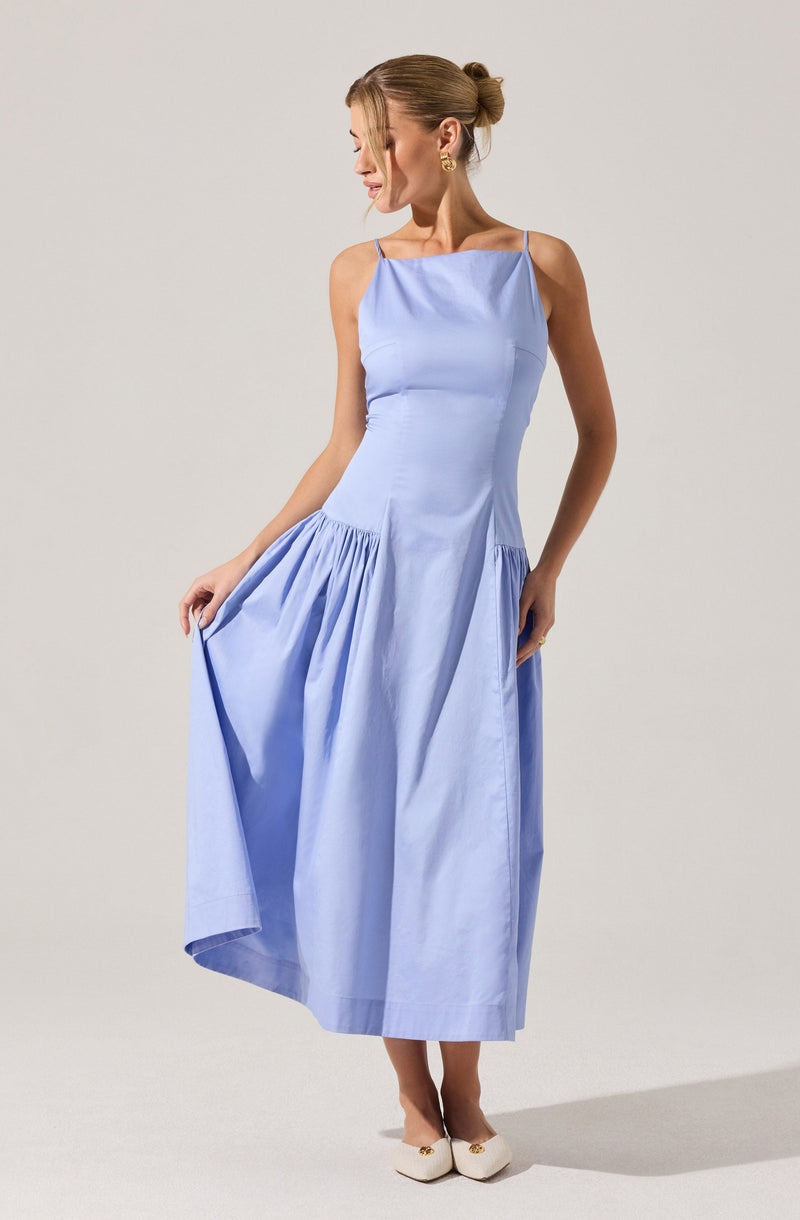 A woman is wearing a light blue sleeveless dress with thin spaghetti straps. The dress has a fitted bodice and a gathered skirt that she is holding up slightly with her left hand. She is also wearing cream-colored shoes with embellishments at the front. Her hair is styled in a neat, twisted bun.