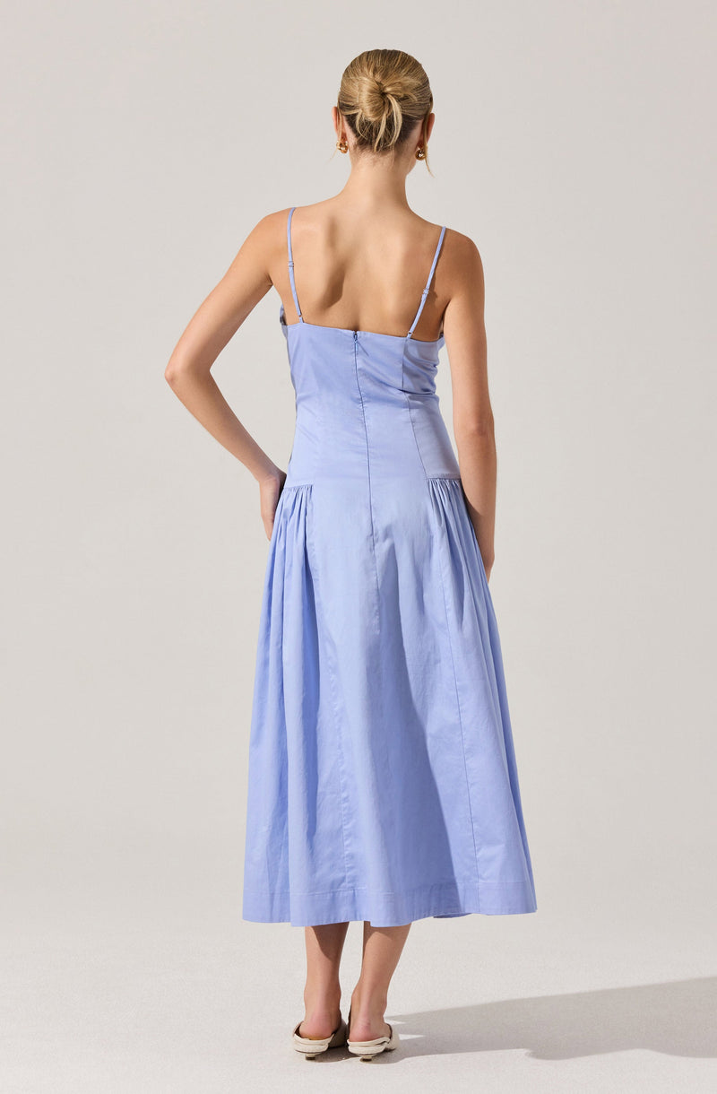 A woman is wearing a light blue, sleeveless dress with thin spaghetti straps. The bodice is fitted, and the skirt is full and gathered, reaching mid-calf length. She is wearing beige, open-toed shoes.