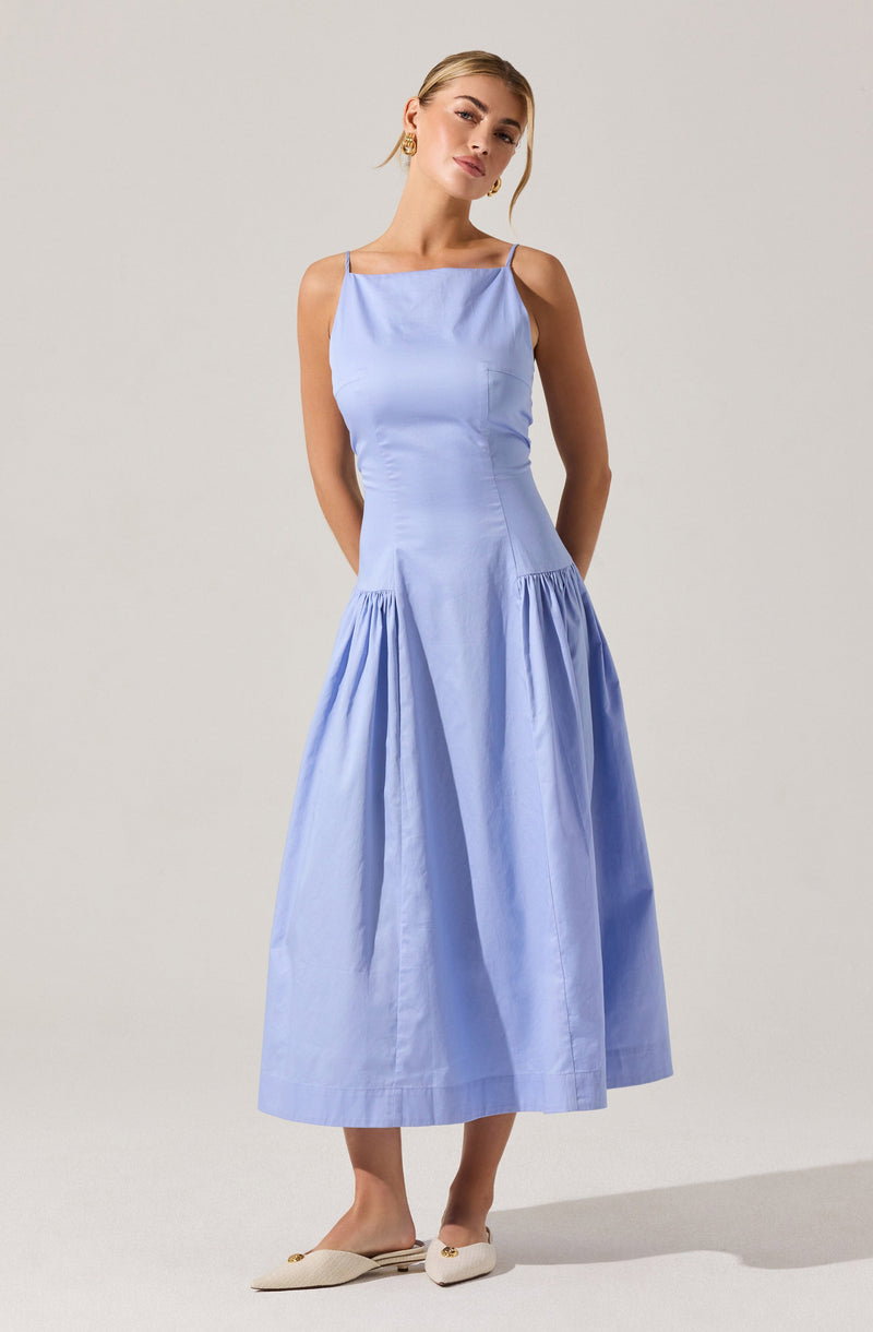 A woman is wearing a sleeveless, light blue dress with thin spaghetti straps. The dress has a fitted bodice and a gathered, full skirt that falls to mid-calf length. She is also wearing cream-colored flat shoes with a bow detail.
