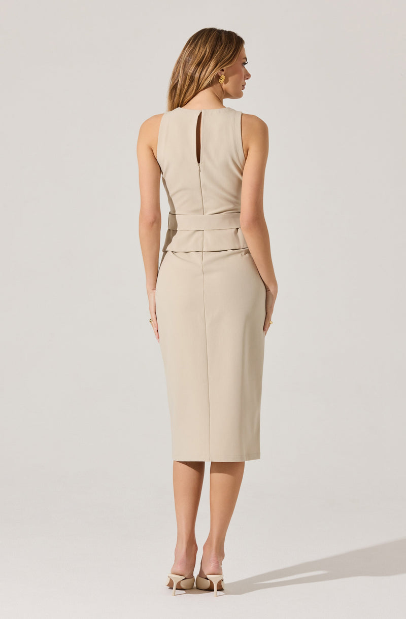 A woman is wearing a sleeveless, beige, knee-length dress with a fitted waist. The dress features a small vertical slit at the back neckline and has a peplum detail at the waist. She is also wearing beige high-heeled shoes.
