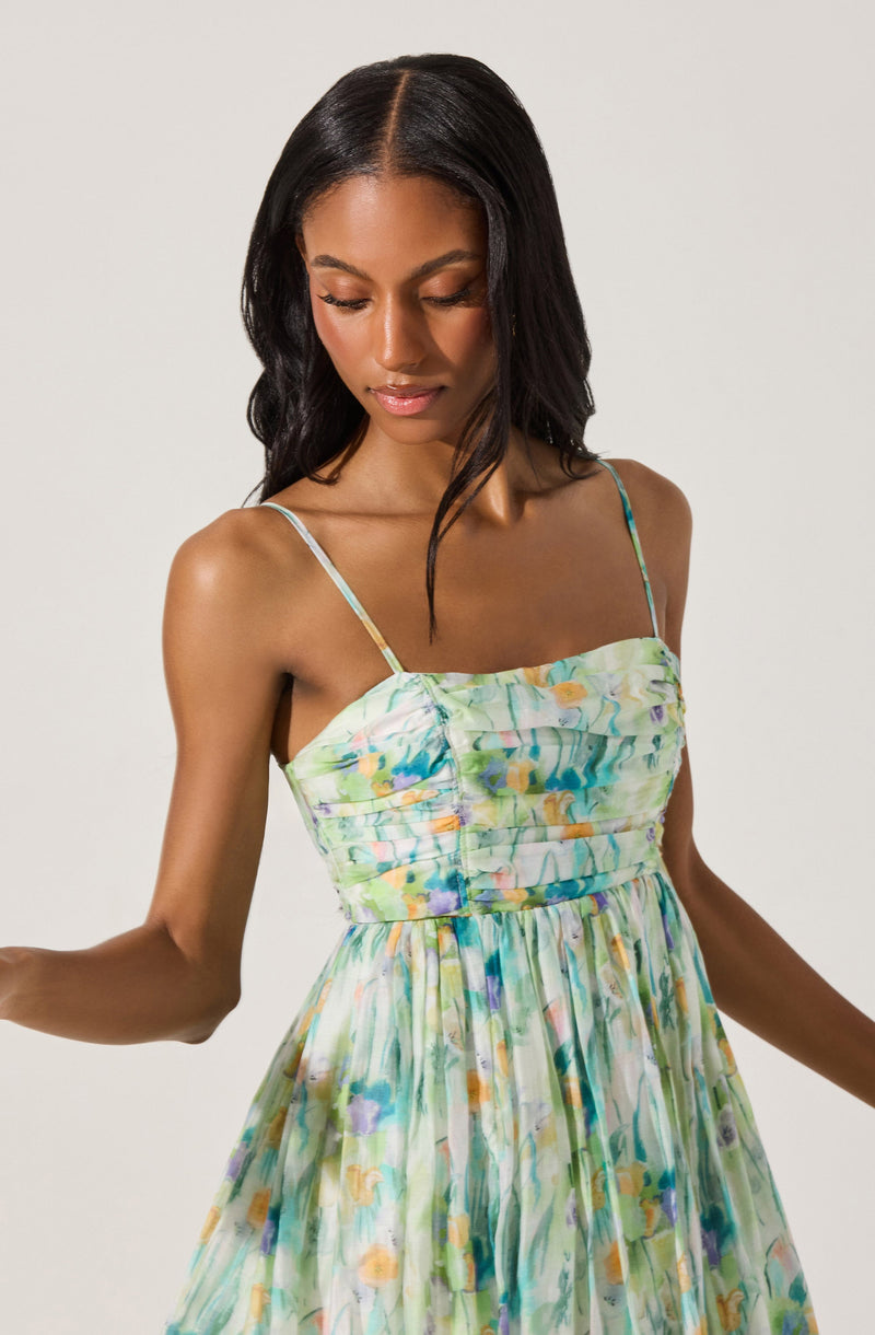 A woman is wearing a sleeveless, summer dress with thin spaghetti straps. The fabric features a colorful, abstract floral pattern in shades of green, blue, orange, purple, and yellow. The bodice of the dress is fitted with subtle ruching, and the skirt is gathered and flows loosely.