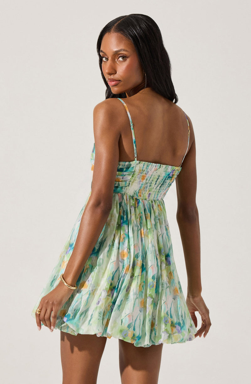 A woman is wearing a sleeveless dress with spaghetti straps. The dress has a fitted, smocked bodice and a full, flared skirt. The fabric features a colorful, abstract pattern with shades of green, blue, orange, and purple.