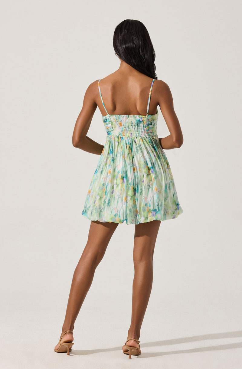 A woman is wearing a sleeveless, short dress with spaghetti straps. The dress features a colorful, abstract watercolor pattern in shades of green, blue, yellow, and hints of purple, with a smocked bodice and a slightly gathered skirt. She is also wearing beige high-heeled sandals.
