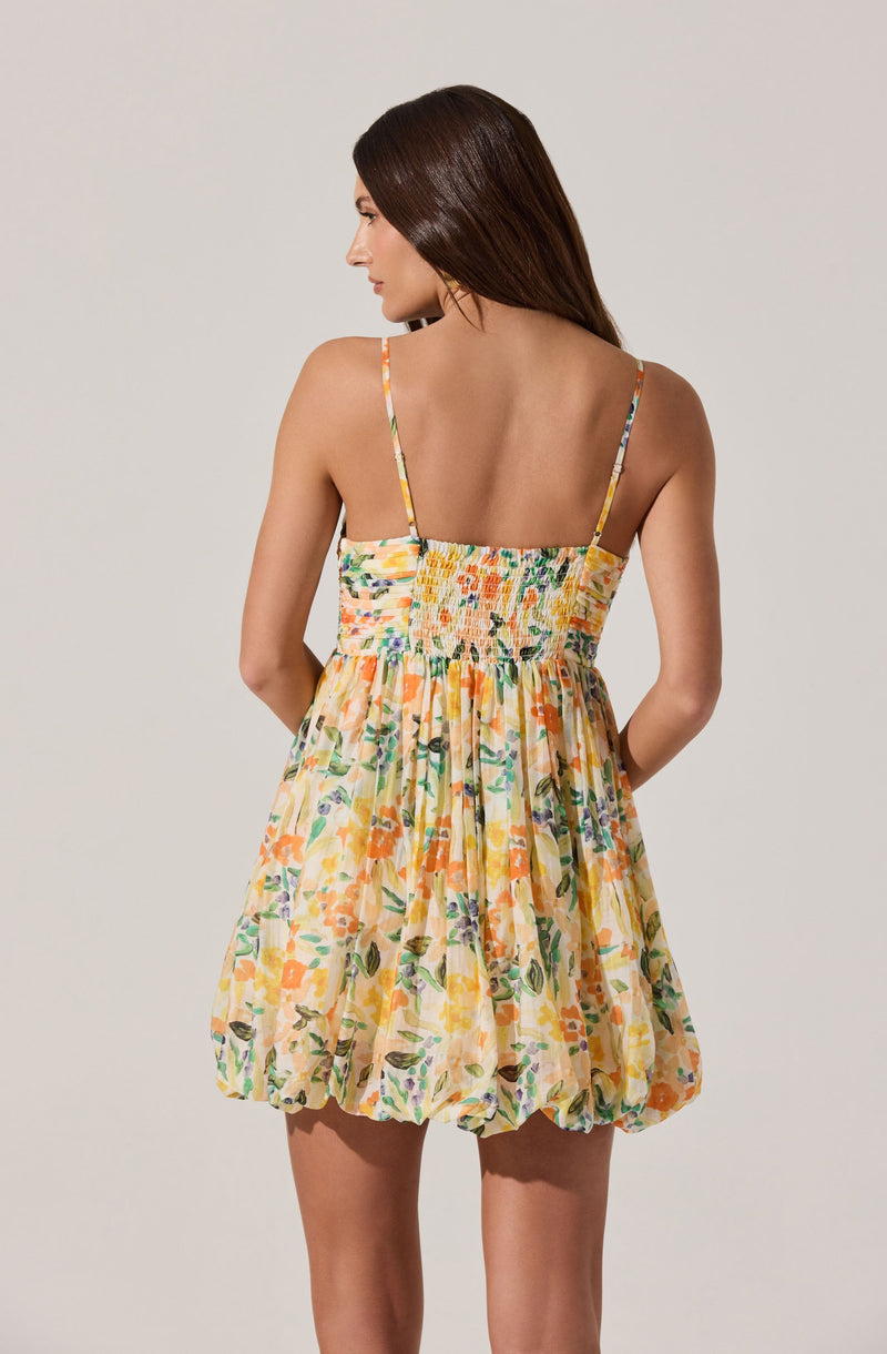 A woman is wearing a sleeveless dress with thin spaghetti straps. The dress has a smocked bodice and a full, gathered skirt. The fabric features a colorful floral print with shades of orange, yellow, green, and purple.