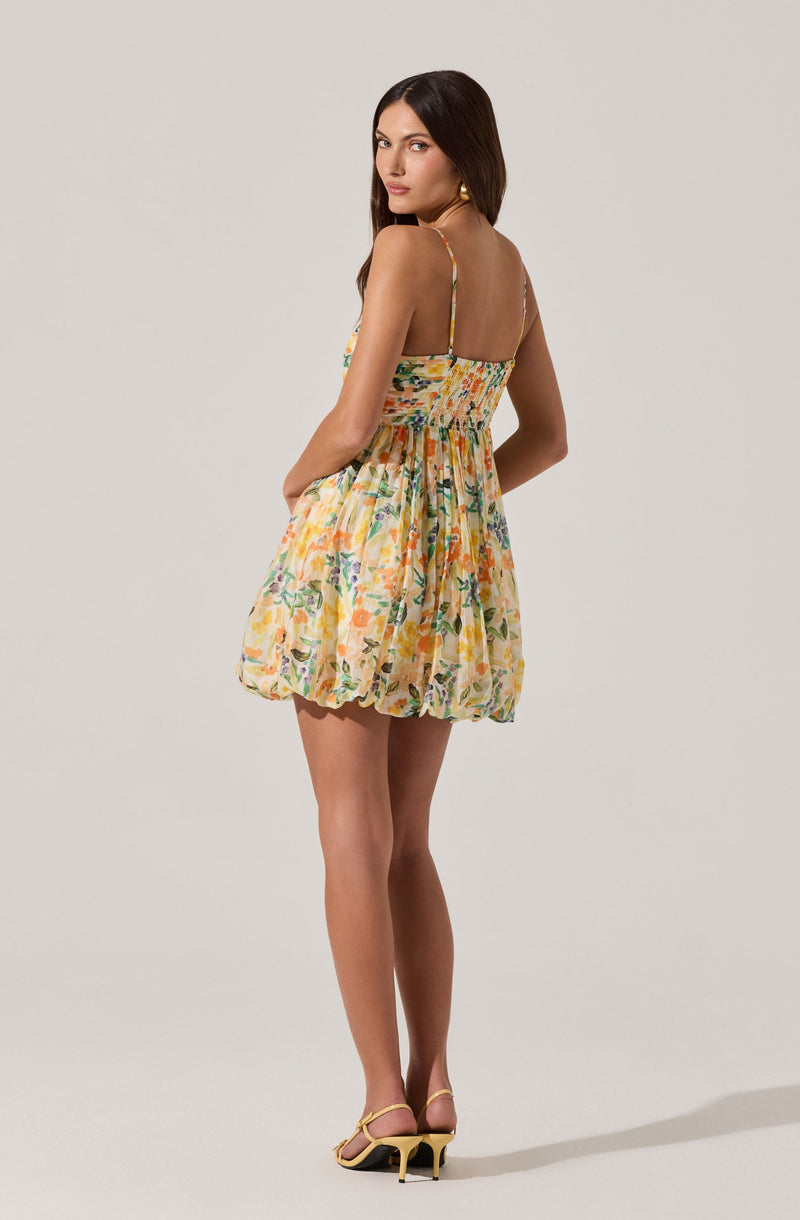 The dress is a sleeveless, knee-length dress with spaghetti straps. It features a floral pattern with shades of yellow, green, purple, and orange on a light, possibly beige or off-white background. The dress has a gathered, elasticized waist and a full, slightly puffy skirt. The model is also wearing a pair of light-colored, open-toe high-heeled shoes with ankle straps.