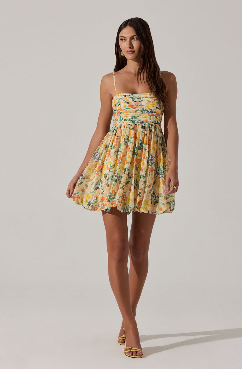 A woman is wearing a sleeveless, floral dress with thin spaghetti straps. The dress features a fitted bodice with horizontal smocking and a gathered, voluminous skirt that falls above the knees. The fabric has a colorful pattern with shades of yellow, orange, green, blue, and purple, depicting various flowers and leaves.