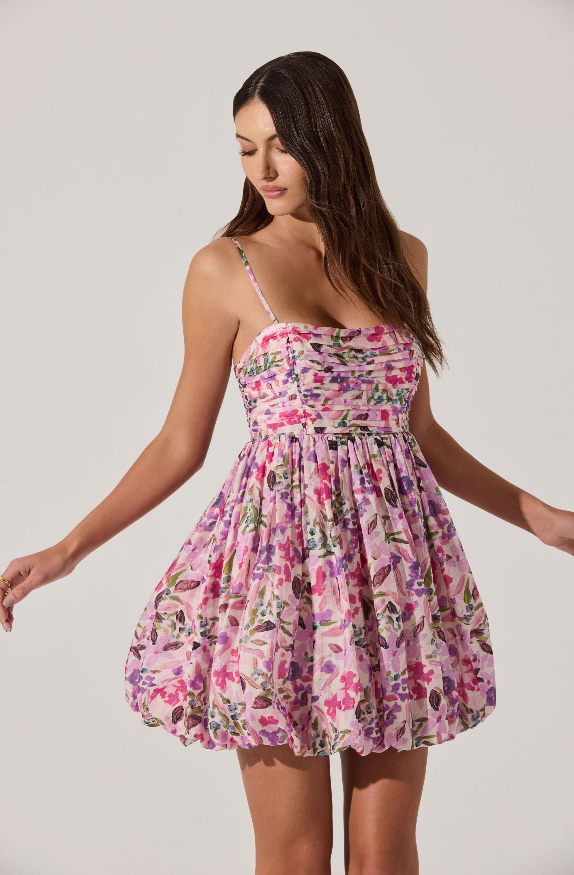 A woman is wearing a sleeveless dress with thin spaghetti straps. The dress has a fitted bodice with a ruched design and a gathered, voluminous skirt. The fabric features a colorful floral pattern with shades of pink, purple, green, and brown.