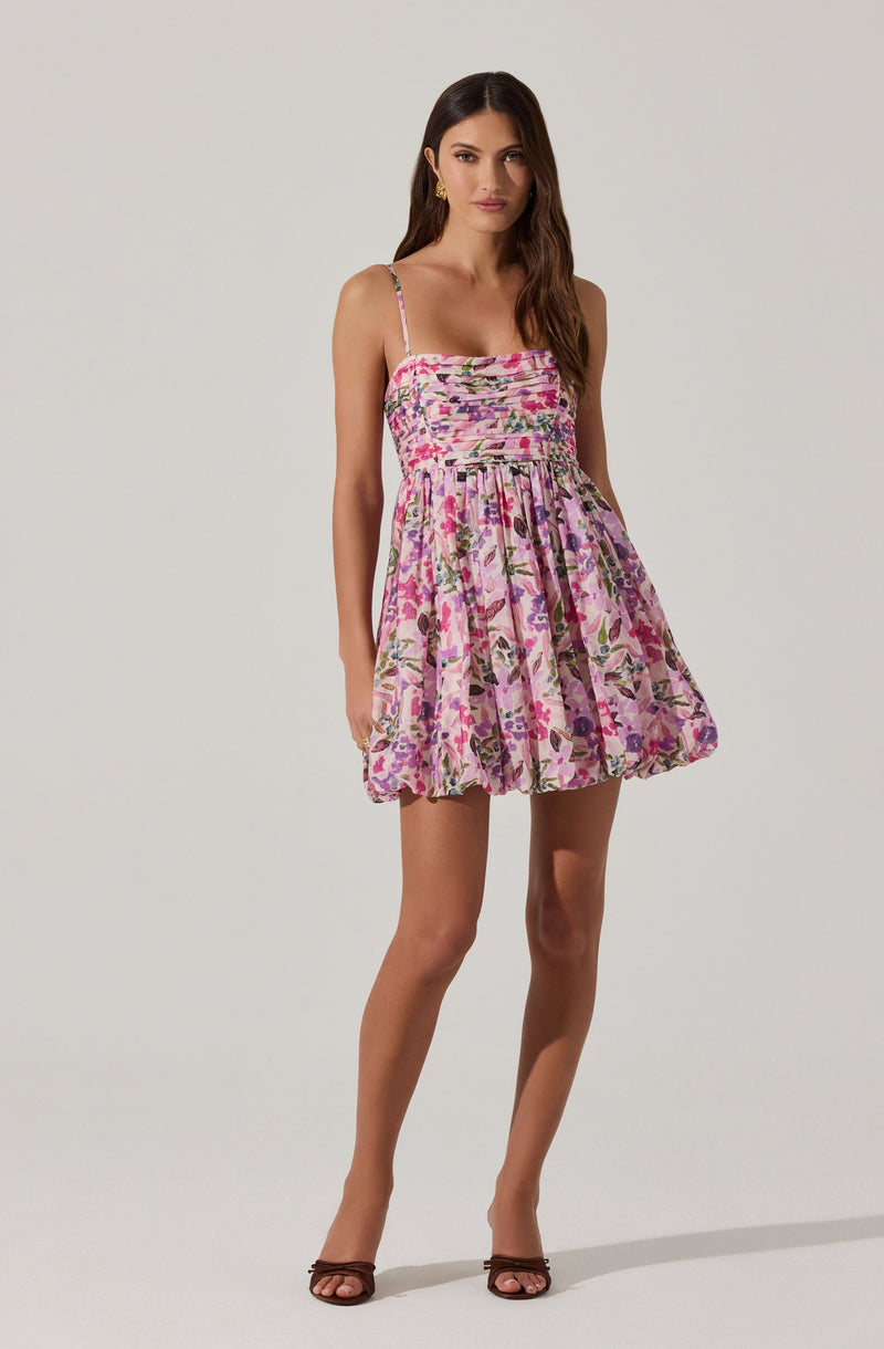 A woman is wearing a sleeveless, above-the-knee dress with spaghetti straps. The dress features a fitted, smocked bodice with horizontal ruched detailing and a full, gathered skirt. The fabric has a floral pattern with shades of pink, purple, green, and beige. She is also wearing open-toed, dark brown high-heeled sandals and gold earrings.