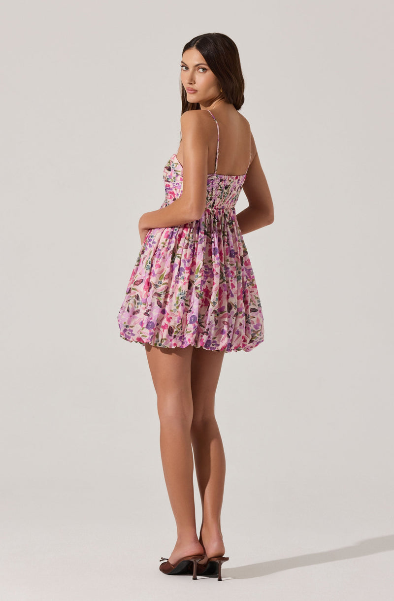 A woman is wearing a short, sleeveless dress with thin spaghetti straps. The dress features a colorful floral print with shades of pink, purple, and green. The bodice is fitted with a smocked detail, and the skirt is voluminous and gathered, creating a bubble hem. She is also wearing a pair of open-toed high-heeled sandals.