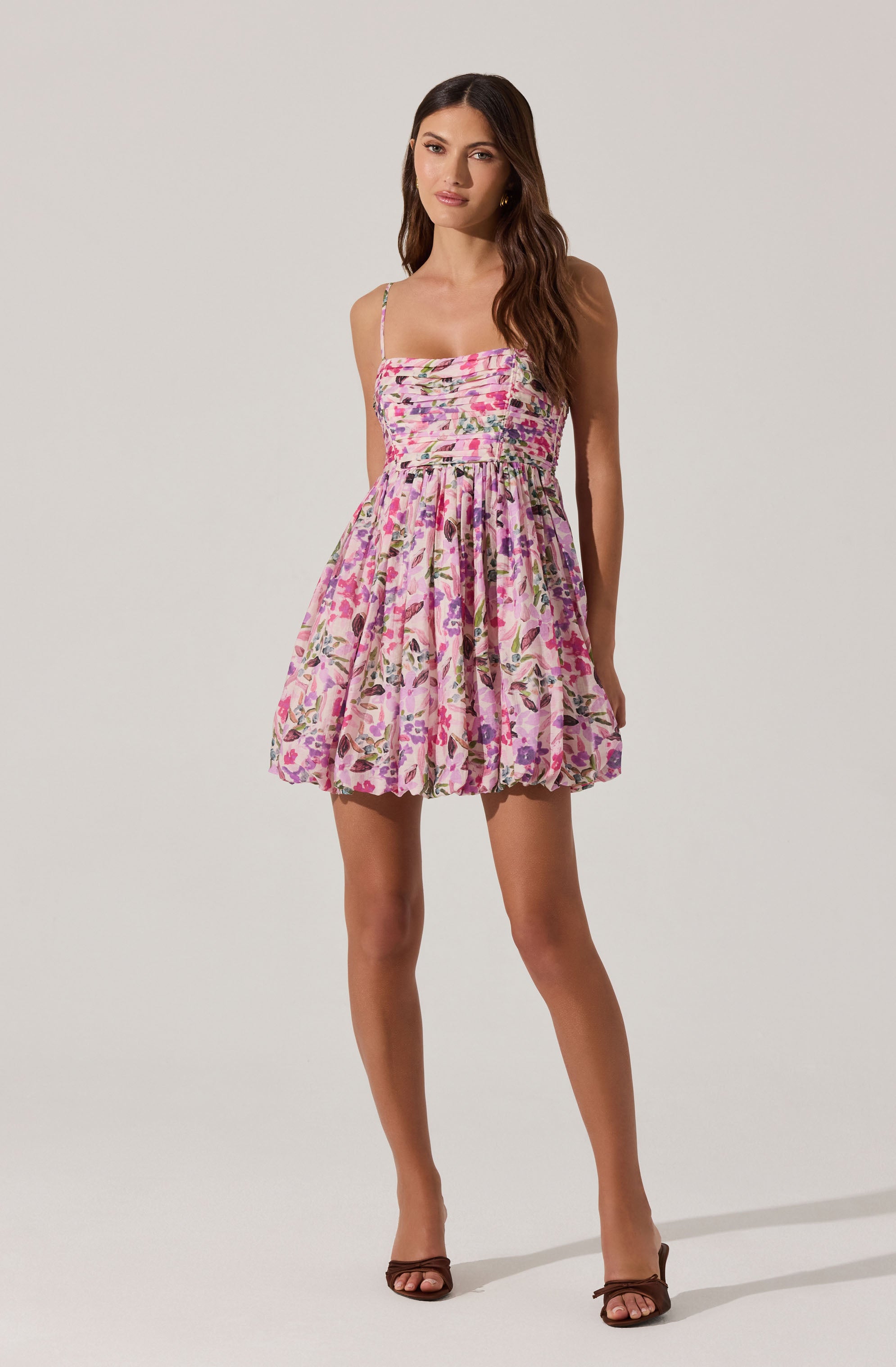 A woman is wearing a sleeveless dress with thin spaghetti straps and a fitted bodice. The dress features a pink floral pattern with various shades of purple, green, and white, and has a gathered, puffed skirt that falls above the knees. She is also wearing open-toed high heels.