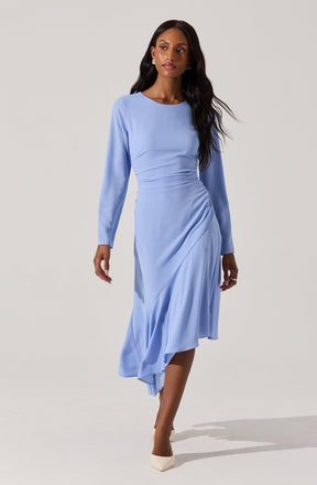 A woman is wearing a light blue, long-sleeved, midi-length dress with a fitted waist and a draped, asymmetrical skirt. The dress has a round neckline and appears to be made of a smooth, flowy fabric. She is also wearing white heels.
