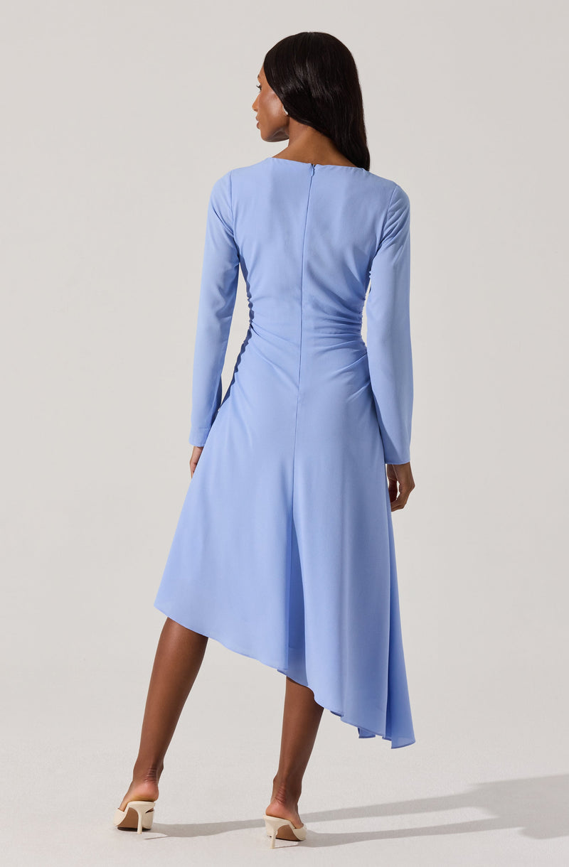 A woman is wearing a light blue, long-sleeved dress with a fitted waist and an asymmetrical hemline that is shorter in the front and longer in the back. The dress appears to be made of a smooth, slightly shiny fabric. She is also wearing beige high-heeled shoes.
