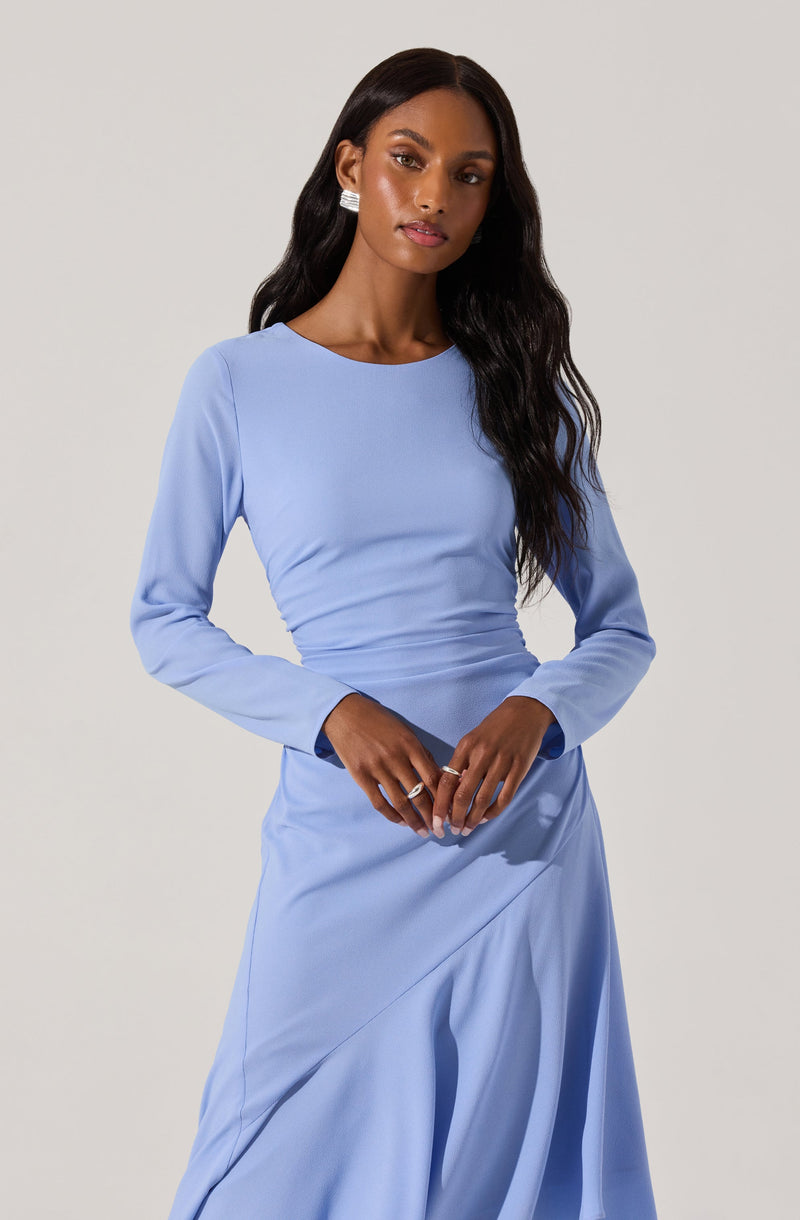 A woman is wearing a light blue long-sleeve dress with a fitted bodice and a flowing skirt. The dress has a round neckline and features subtle ruching at the waist. She is accessorized with large, rectangular statement earrings and a ring on her right hand.