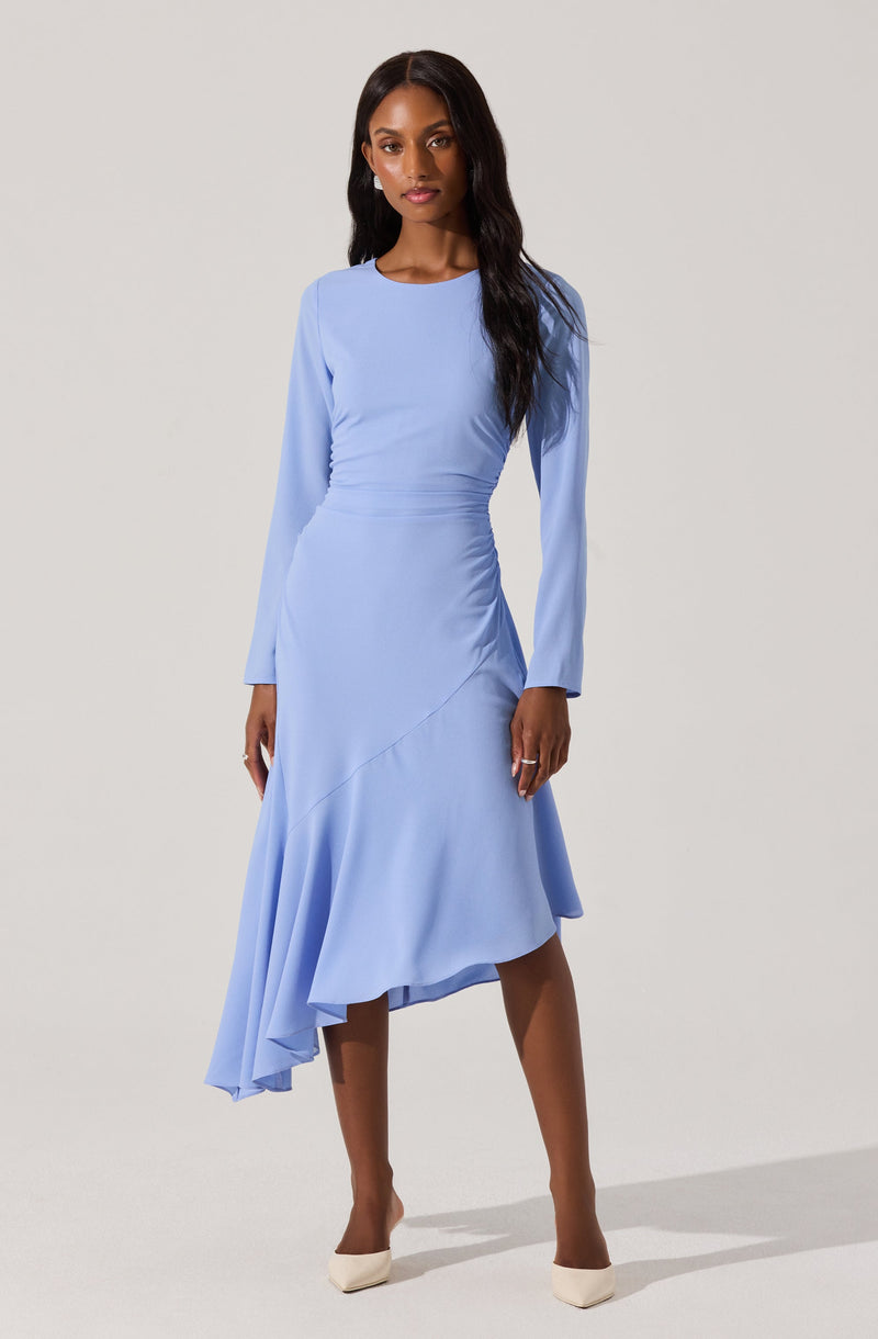 A woman is wearing a light blue, long-sleeved dress with a fitted bodice and a flowing, asymmetrical skirt that is longer on one side. The dress has a round neckline and a smooth, elegant fabric. She is also wearing nude-colored high heels.