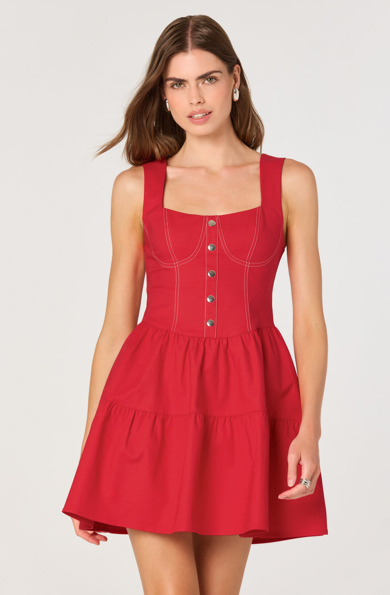 A woman is wearing a sleeveless, knee-length red dress with wide shoulder straps. The dress features a fitted bodice with visible white stitching outlining the bodice and a vertical row of metal buttons down the front. The skirt part of the dress is gathered and flares out with a tiered design, creating a ruffled effect.
